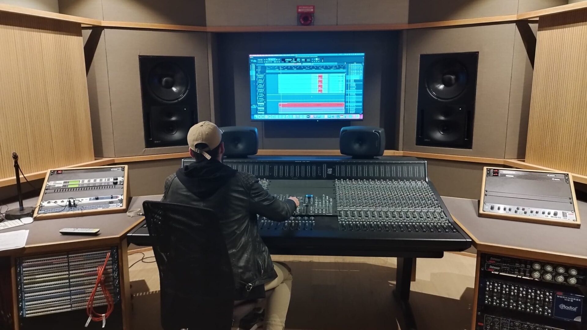 A person sits at a desk in a music studio, surrounded by recording equipment. They are focused on their work, with studio monitors on and a computer screen in front of them.